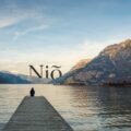 Image d'illustration de Homepage de Niõ - Bougies Fait Main et parfums d'ambiance authentiques : Image représente une personne assise sur un ponton aux abords d'un lac face à des montagnes recouvertes de forêts sous un ciel bleu. Le logo de Niõ se soulève entre les montagnes comme un élément planté dans le décor, le paysage et l'environnement. L'image représente les engagements éco-responsables de Niõ, le soin de l'environnement et la proximité avec les clients.