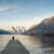 Image d'illustration de Homepage de Niõ - Bougies Fait Main et parfums d'ambiance authentiques : Image représente une personne assise sur un ponton aux abords d'un lac face à des montagnes recouvertes de forêts sous un ciel bleu. Le logo de Niõ se soulève entre les montagnes comme un élément planté dans le décor, le paysage et l'environnement. L'image représente les engagements éco-responsables de Niõ, le soin de l'environnement et la proximité avec les clients.