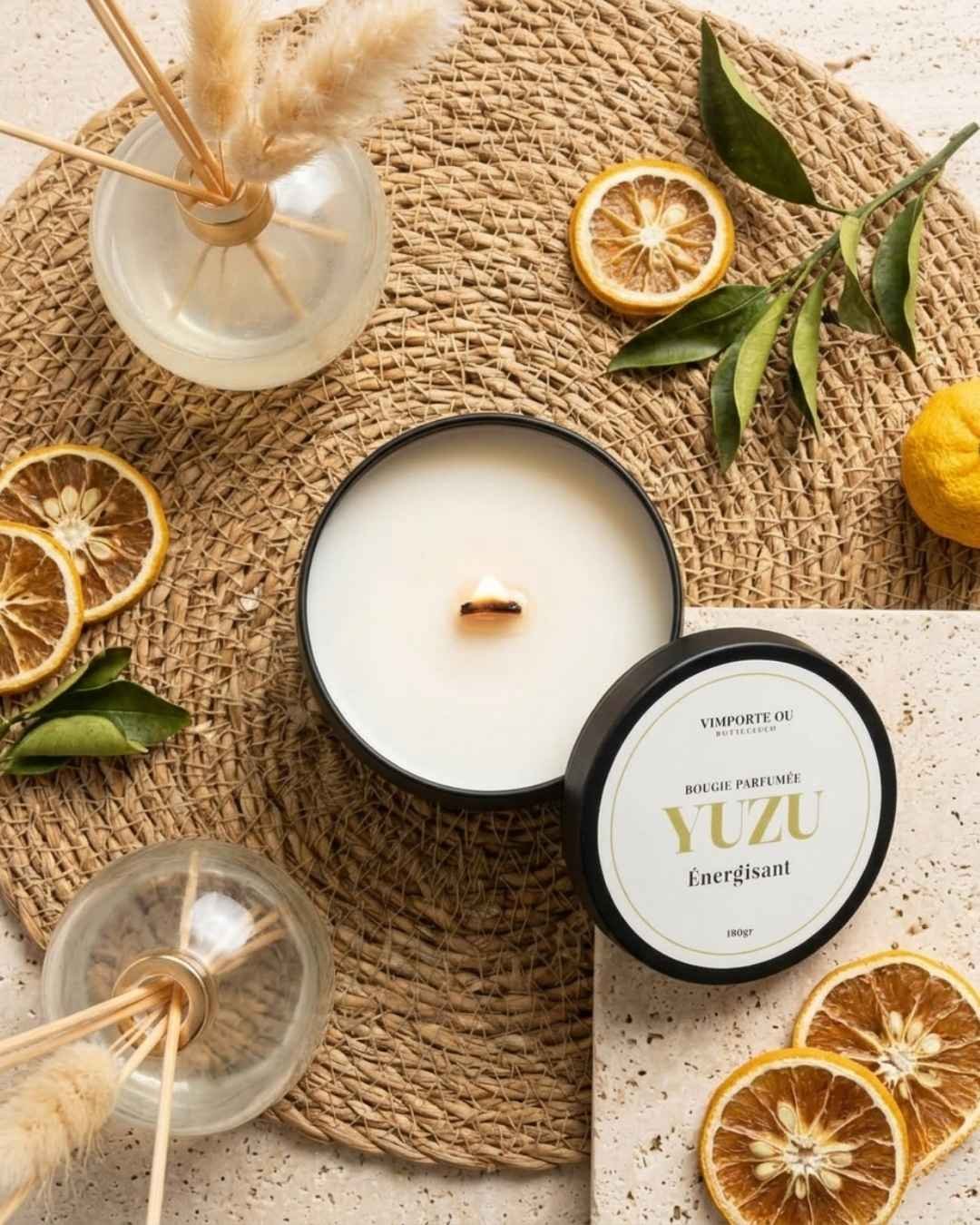 La bougie parfumée naturelle Yuzu énergisant de Niõ représente la famille olfactive hespéridée dans une composition photographique vue du ciel avec la bougie ouverte, et au tour des yuzu frais et des tranches de yuzu séchées, des feuilles de yuzu et un diffuseur de parfum d'ambiance à batônnets 