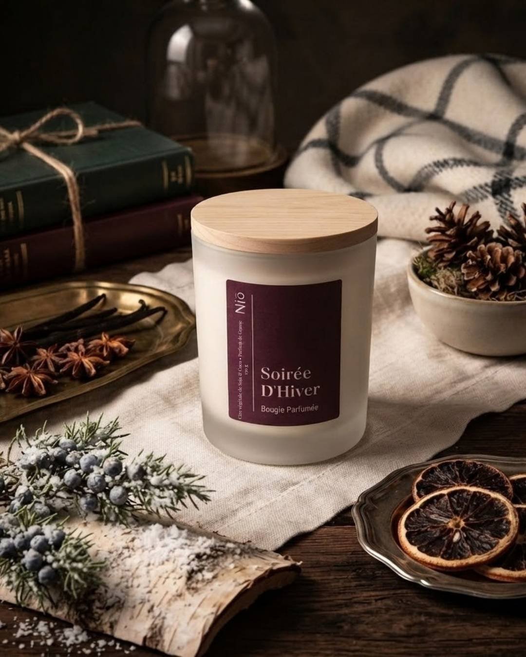 Bougie Soirée d'Hiver en verre givré sur lin crème, entourée d'orange séchée, de pommes de pin et de livres